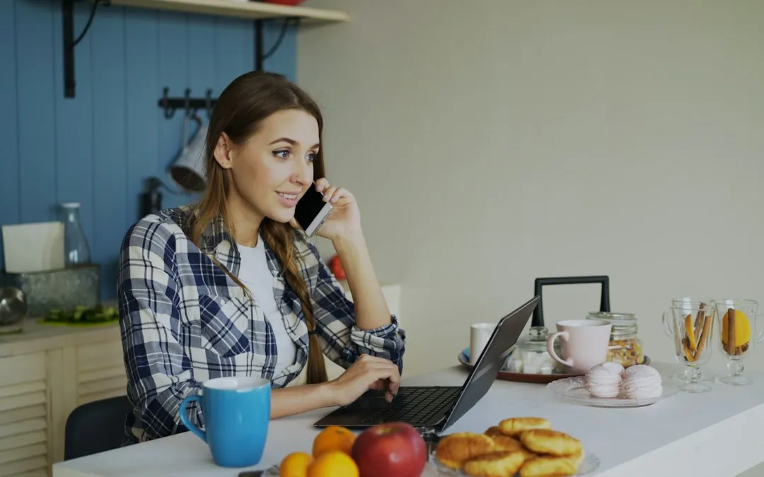 A woman at a kitchen table with a laptop open, talking on the phone.
